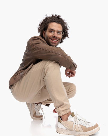 happy young man with glasses and curly hair crouching with elbow on knee in front of white backgroundの写真素材