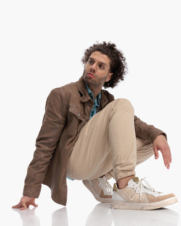 side view of curly hair man with glasses crouching and looking to side while holding elbow on knee on white backgroundの写真素材