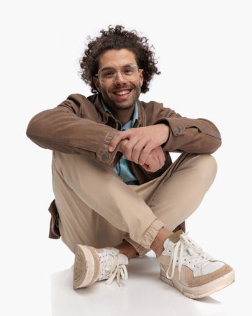 excited casual man with glasses and curly hair laughing and holding hands while elbows are on knees in front of white backgroundの写真素材