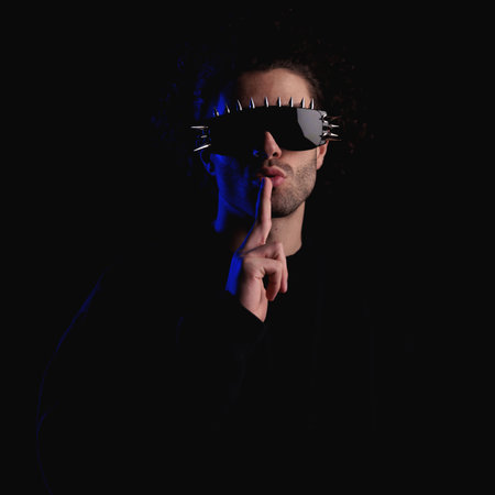 cool fashion man with glasses making silence gesture while hiding from police in front of black background in studioの写真素材