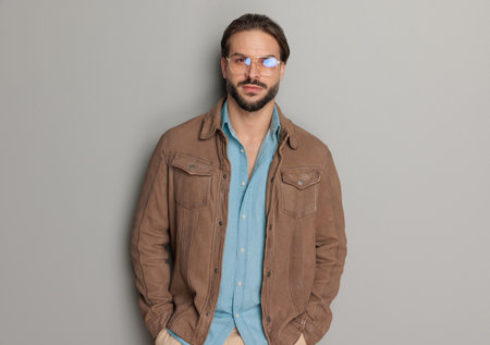 portrait of casual man wearing brown leather jacket and denim shirt, holding hands in pockets and posing in front of light grey backgroundの写真素材