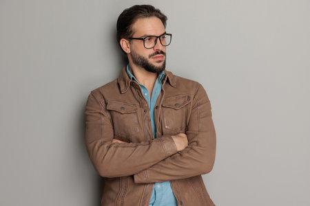portrait of attractive bearded guy with glasses folding arms and looking to side while posing in front of grey background in studioの写真素材