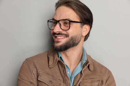 portrait of happy casual man wearing brown leather jacket and denim shirt, looking to side and smiling in front of light grey backgroundの写真素材