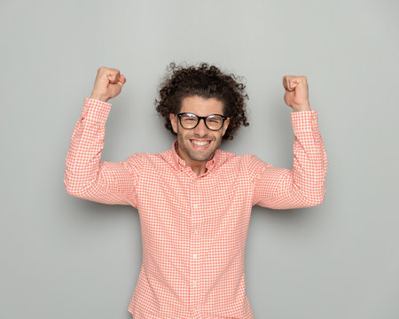 portrait of proud young man with glasses holding fists up and celebrating victory while posing in front of grey backgroundの写真素材