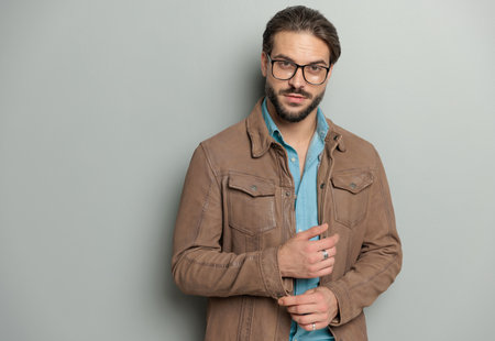 attractive man with glasses in brown leather jacket adjusting sleevess and looking forward while posing in front of grey backgroundの写真素材