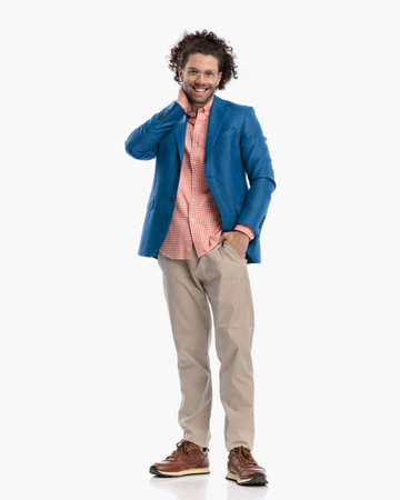 full body picture of happy casual young man with curly hair with hand in pocket touching neck and laughing while standing in front of white background in studioの写真素材