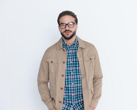 sexy casual man with glasses in plaid shirt and jacket holding hands in pockets and posing in front of grey backgroundの写真素材