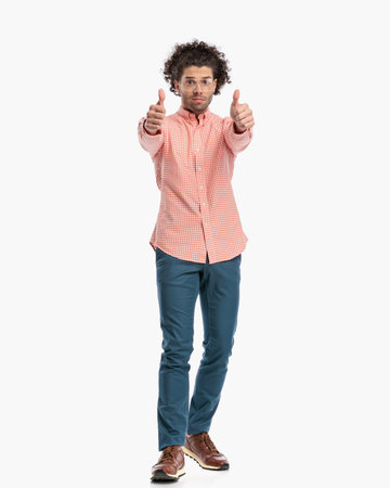 attractive fashion man with curly hair making thumbs up gesture and posing while walking in front of white background in studioの写真素材