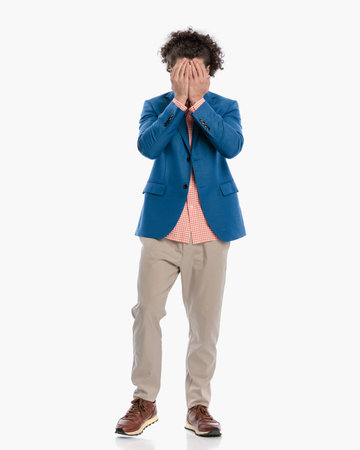 shy young man with curly hair covering face and hiding behind while standing in front of white background in studioの写真素材