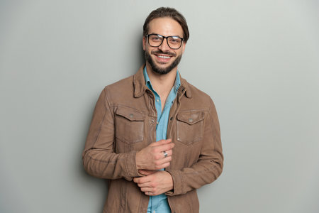 happy casual man with glasses wearing brown leather jacket and adjusting sleeves, smiling and posing in front of grey backgroundの写真素材