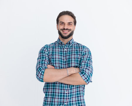 portrait of handsome young man with beard in plaid shirt crossing arms and smiling in front of grey background in studioの写真素材