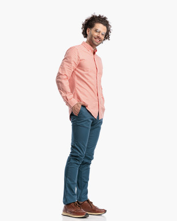 side view of smart casual guy with glasses holding hands in pockets and smiling while posing in front of white background in studioの写真素材