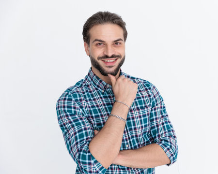 happy young man in plaid shirt folding arms and touching chin while smiling in front of grey background in studioの写真素材