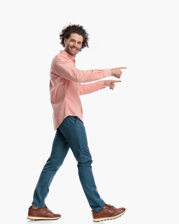 side view of happy curly hair man smiling and pointing fingers while walking in front of white background in studioの写真素材