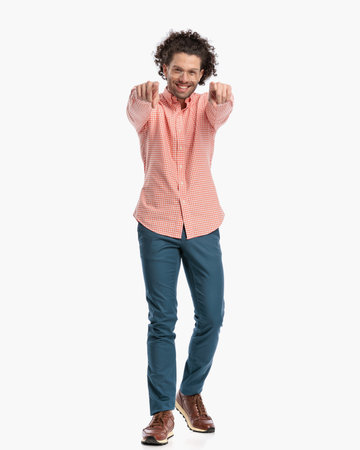happy young man with glasses pointing fingers forward and smiling while walking in front of white background in studioの写真素材