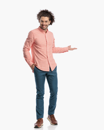 happy cute man with curly hair wearing glasses, holding one hand in pocket and showing and presenting to side while walking on white backgroundの写真素材