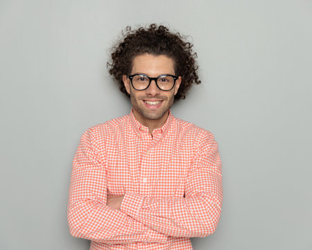 portrait of smart casual man with curly hair and glasses crossing arms and smiling in front of grey background in studioの写真素材