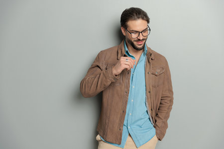 unshaved casual guy with glasses holding hand in pocket and looking down while posing in front of grey backgroundの写真素材