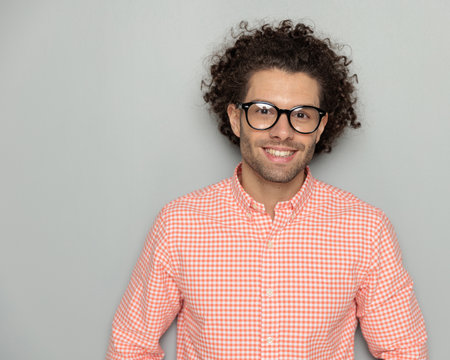 portrait of curly hair man in plaid shirt with glasses smiling and being happy, standing and posing in front of grey background in studioの写真素材