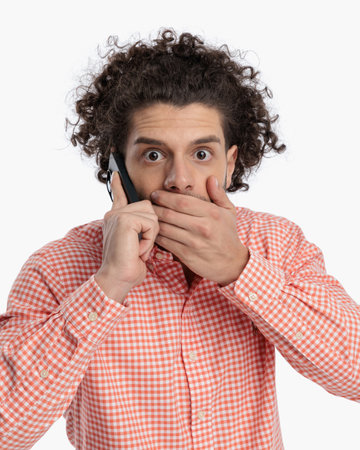 portrait of shocked man covering mouth with hand while talking on the phone in front of white backgroundの写真素材