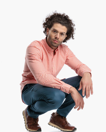 sexy gashion man with glasses and curly hair crouching and holding arms on knees in front of white backgroundの写真素材