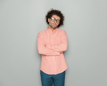 tired young man with curly hair crossing arms and posing with eyes closed in front of grey backgroundの写真素材