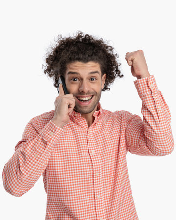 excited young man with curly hair talking on the phone and celebrating with fists up, laughind and being happy on white backgroundの写真素材