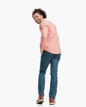 back view picture of happy young man looking behind and smiling while walking with hand in pocket in front of white background in studioの写真素材