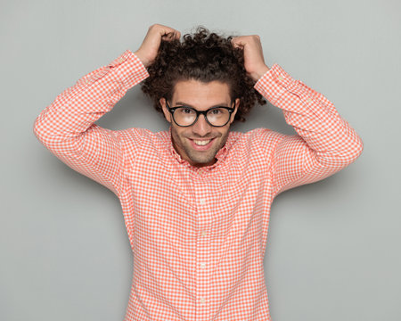 portrait of happy young man with glasses pulling curly hair and smiling while posing on grey background in studioの写真素材