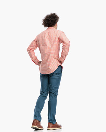 casual young guy holding hands on hips and looking away while walking in front of white background in studioの写真素材