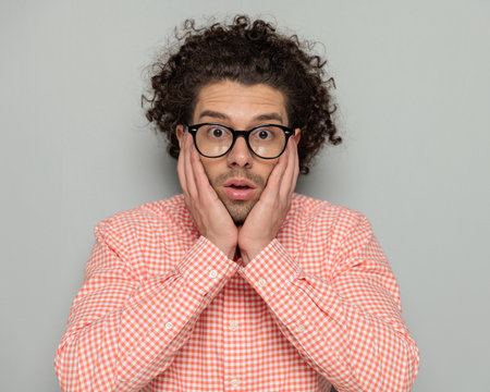 surprised attractive guy with glasses holding palms on face and opening mouth in a shocked manner on grey backgroundの写真素材