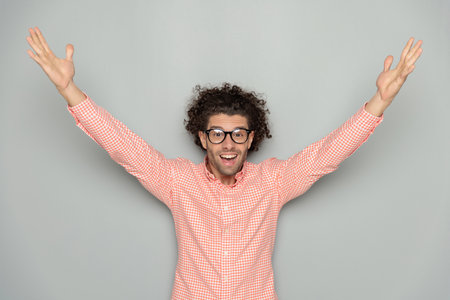 excited young man with curly hiar and eyeglasses holding arms in the air and cheering while posing in front of grey backgroundの写真素材