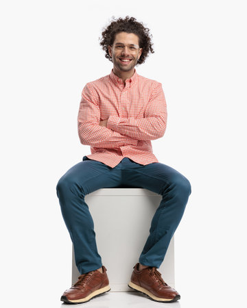 excited young man with curly hair and glasses crossing arms, sitting and laughing in front of white background in studioの写真素材