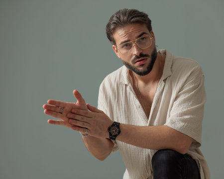 handsome guy with glasses holding elbow on knee and touching palms while posing in front of grey...