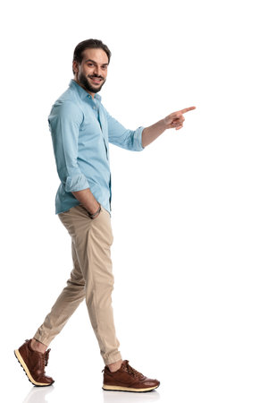 side view of casual man pointing finger forward and walking with hand in pocket, being happy and smiling on white backgroundの写真素材