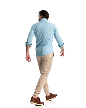 rear view of casual man wearing blue denim shirt with chino pants and sneakers walking and looking to side in front of white backgroundの写真素材