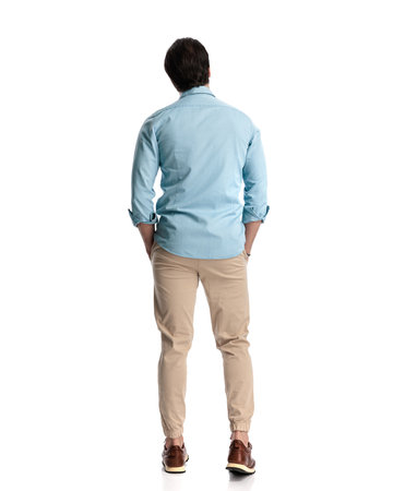 back view of casual man holding hands in pockets and standing in front of white background in studioの写真素材