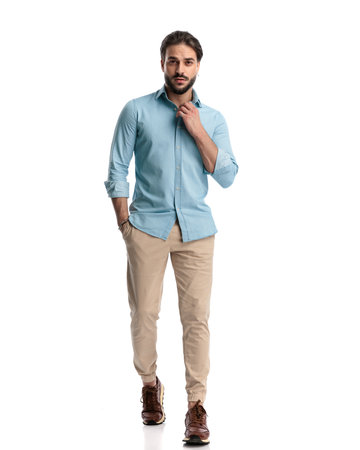 sexy unshaved young man walking with hand in pocket and adjusting shirt in front of white backgroundの写真素材