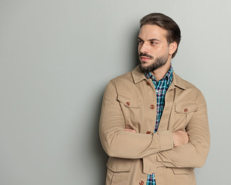 confident young man in beige jacket crossing arms and looking to side while posing in front of grey backgroundの写真素材