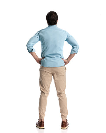 back view of casual young man holding hands on hips and looking forward in front of white backgroundの写真素材