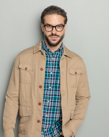 cool casual young man with glasses holding hand in pocket, looking forward and confidently posing on grey backgroundの写真素材