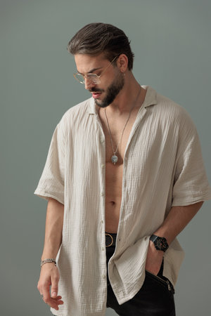 confident bearded man with glasses holding hand in pocket and looking down in front of grey backgroundの写真素材