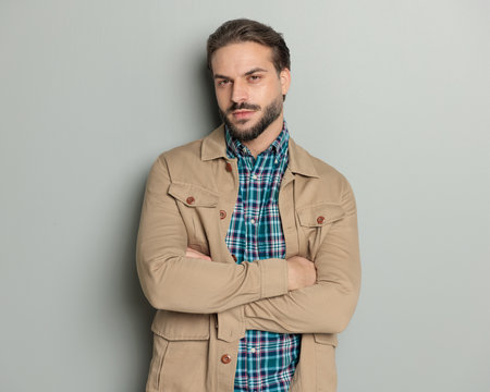 handsome bearded casual man in jacket crossing arms and looking forward in front of grey backgroundの写真素材