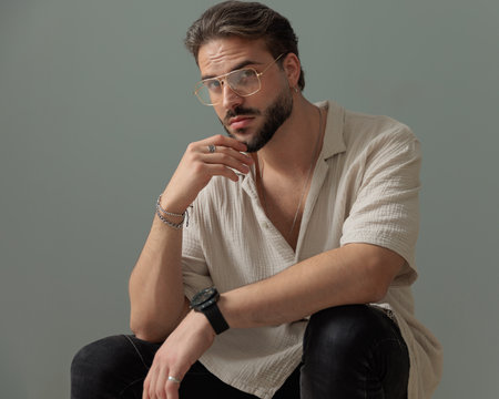 cool bearded guy with glasses holding elbows on knees and looking forward while thinking in front of grey backgroundの写真素材
