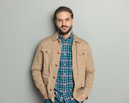 portrait of handsome casual man in beige jacket with plaid shirt holding hands in pockets and posing on grey backgroundの写真素材