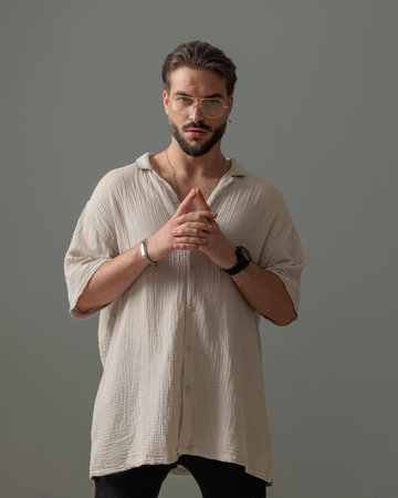 attractive young man with glasses looking forward and holding palms in fashion pose in front of grey backgroundの写真素材