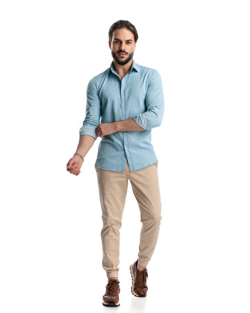 full body picture of sexy casual young man with beard adjusting sleeve and walking in front of white background in studioの写真素材
