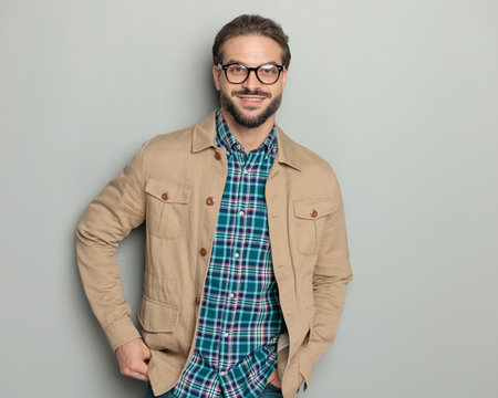 happy casual man with glasses holding hands in pockets and smiling in front of grey backgroundの写真素材