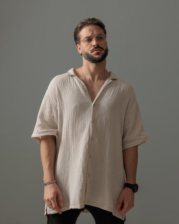 sexy bearded man with glasses looking away and posing in a fashion way in front of grey background in studioの写真素材