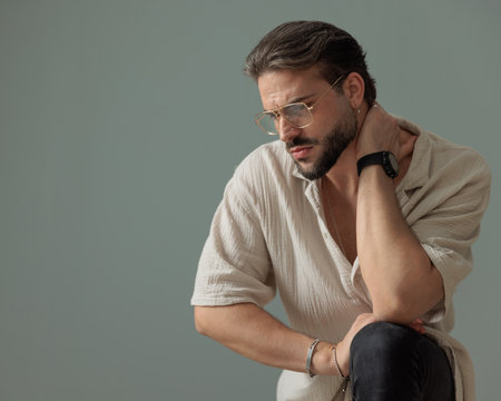 cool bearded man with glasses posing with hand behind neck and looking away posing with knee up in front of grey backgroundの写真素材